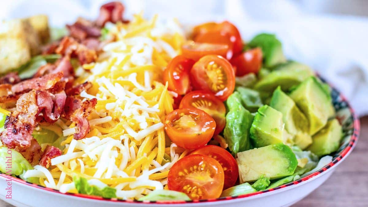 A wide image showing a close-up of BLT salad with bacon, shredded cheese, cherry tomatoes, avocado, and lettuce in a white bowl with a red rim.