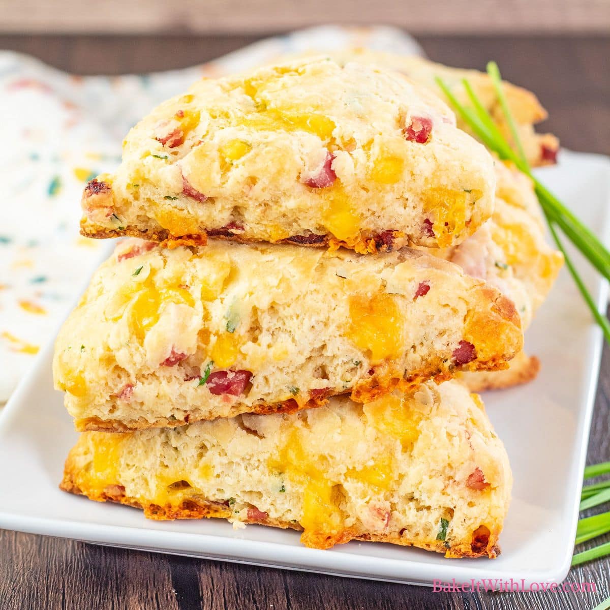 A square image a stack of three golden homemade wedge-shaped scones studded with diced ham, chives, and cheese on a white square plate.