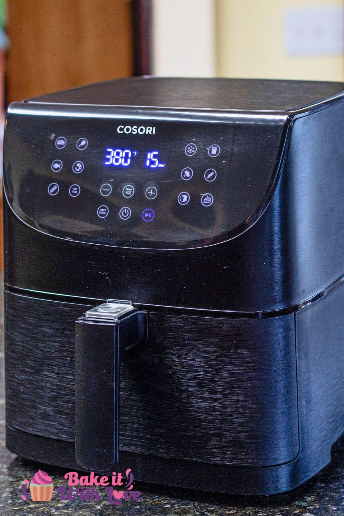 A tall image shows a black Cosori air fryer placed on a kitchen counter. The air fryer features a digital control panel on the front with various touch-sensitive buttons and a bright blue LED display indicating a temperature of 380°F and a timer set for 15 minutes. The lower right of the image includes the "Bake it With Love" logo with a cupcake.