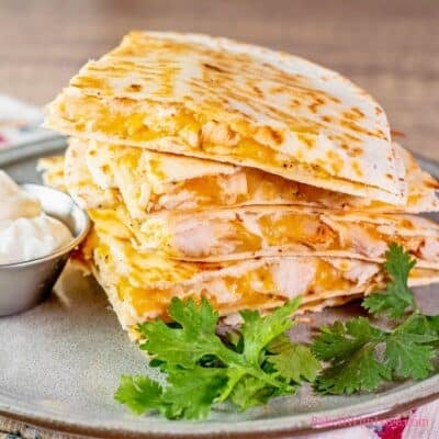 A square image showing crispy quesadilla wedges stuffed with rotisserie chicken and melted cheese, plated with sour cream and garnished with fresh cilantro leaves.