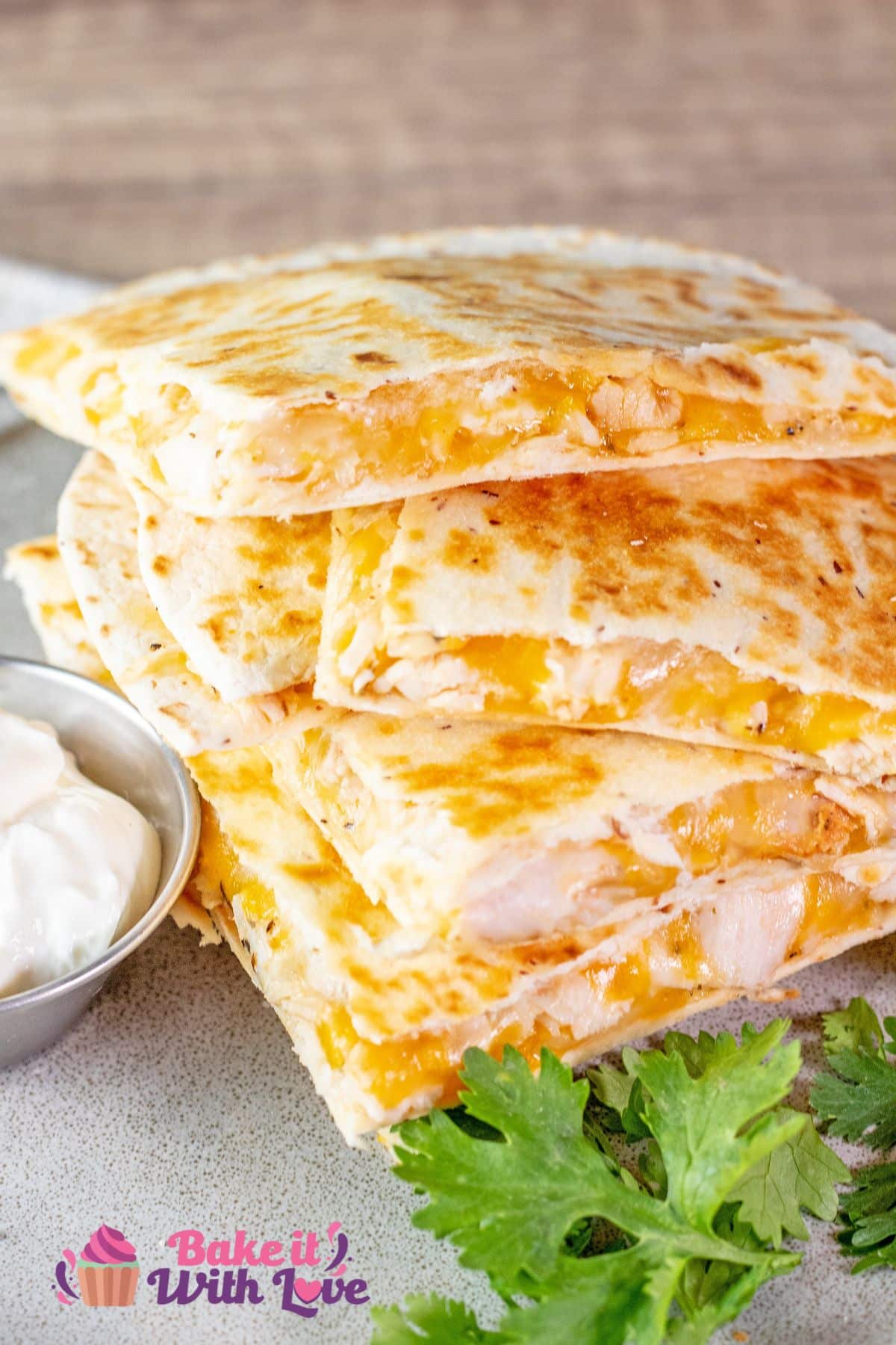 A tall image showing a stack of golden, crispy rotisserie chicken quesadillas loaded with melted cheddar cheese, served with a side of sour cream and fresh cilantro with the BakeItWithLove logo in the bottom left corner.