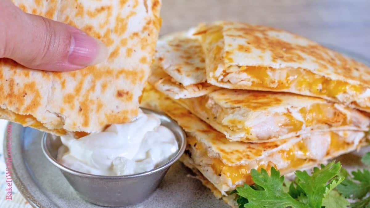 A wide image showing a hand dipping a cheesy slice of rotisserie chicken quesadilla into creamy sour cream, with more quesadilla wedges stacked behind it on the plate.