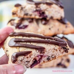 Cherry chocolate chip scones stacked in background with a wedge sliced in half and the open cut side lifted by hand in front of them.