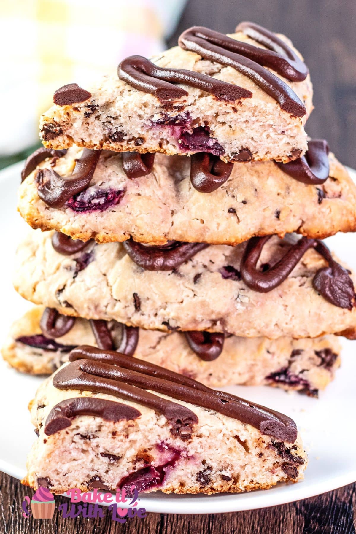 Stacked baked cherry chocolate chip scones with drizzled chocolate ganache topping on white plate with dark wooden background.