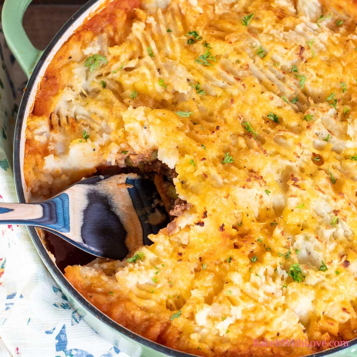 A square image of a golden baked hamburger pie casserole topped with cheesy mashed potatoes with a spoon scooping out a serving.