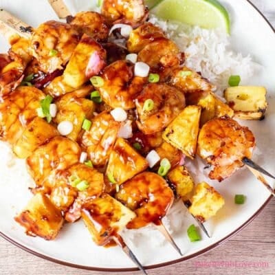 A square image showing 4 grilled pineapple and shrimp skewers coated in sticky caramelized sauce over white rice with a lime on a white plate.