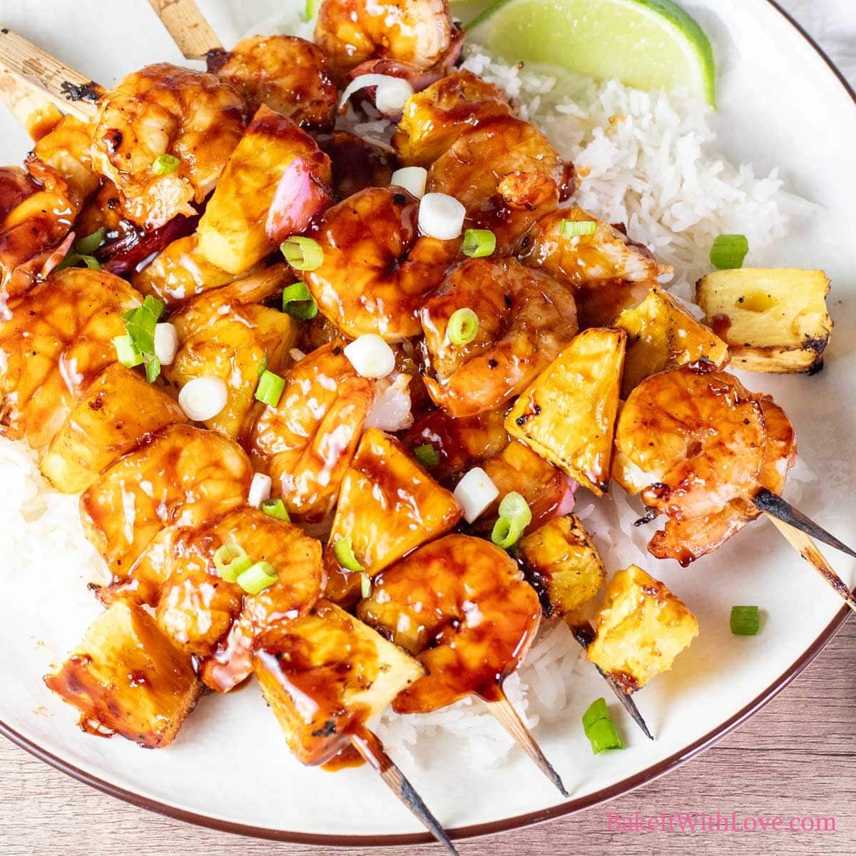 A square image showing 4 grilled pineapple and shrimp skewers coated in sticky caramelized sauce over white rice with a lime on a white plate.